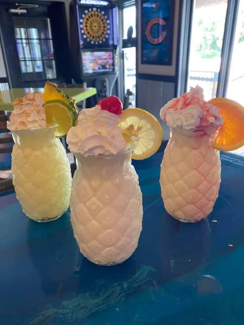 Three colorful tropical drinks in pineapple-shaped glasses, topped with whipped cream and garnished with fruit slices.