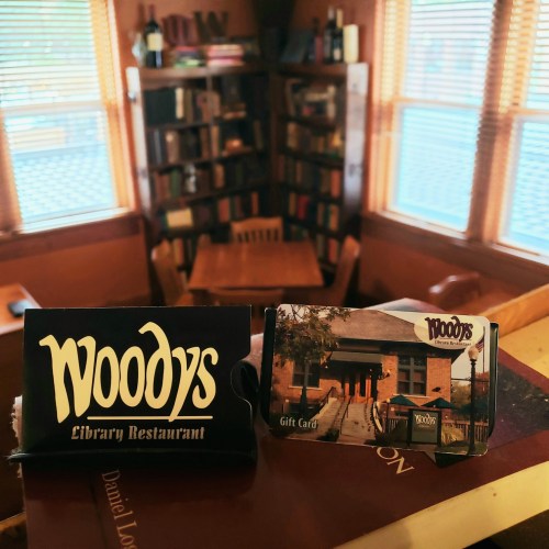 Two Woody's Library Restaurant gift cards on a table, with a cozy library setting in the background.