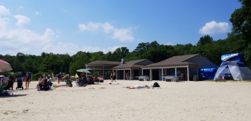 A sandy beach with people relaxing, umbrellas, and a building in the background surrounded by trees under a blue sky.