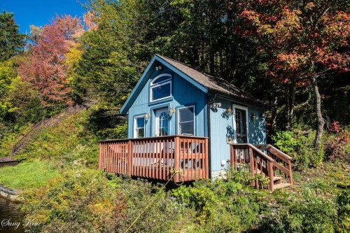 A small blue cabin with a wooden deck surrounded by colorful autumn foliage and trees.