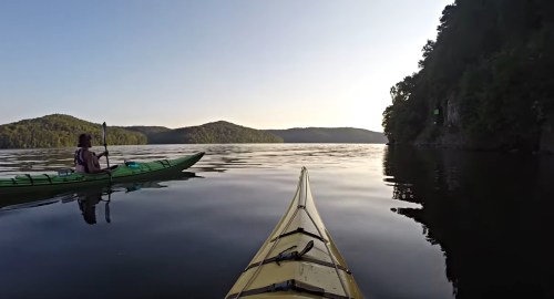 A serene lake scene with two kayakers paddling near lush green hills under a clear sky.