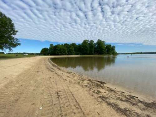 A sandy beach curves along a calm lake, with trees and a blue sky filled with fluffy clouds in the background.