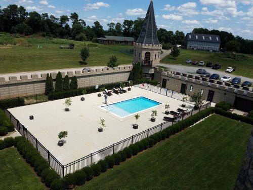 Aerial view of a pool surrounded by lounge chairs, with a castle-like structure and green landscape in the background.