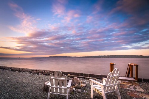 Two white Adirondack chairs facing a calm lake at sunset, with colorful clouds reflecting in the water.