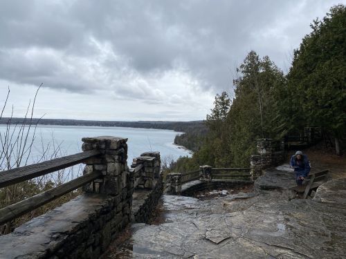 A scenic view of a lake from a stone overlook, with a person sitting on a bench under a cloudy sky.