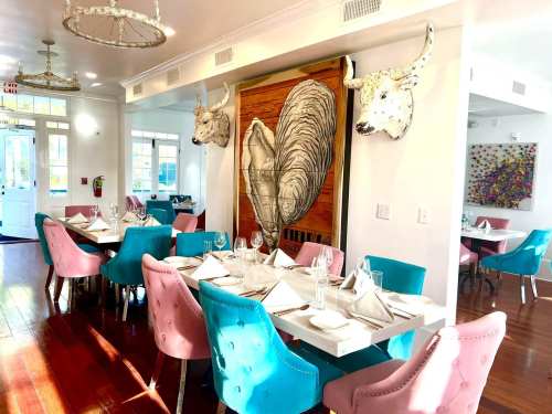 Bright restaurant interior featuring pink and blue chairs, a wooden wall art with bull heads, and elegantly set tables.