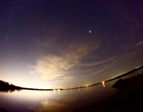 A serene night sky filled with stars reflects on a calm lake, with faint clouds and distant lights along the shore.