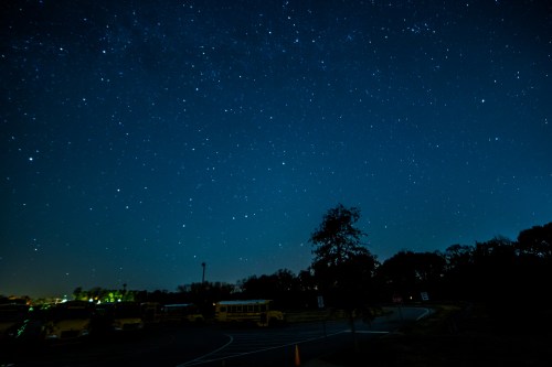 A clear night sky filled with stars above a dark landscape, with silhouettes of trees and parked school buses below.