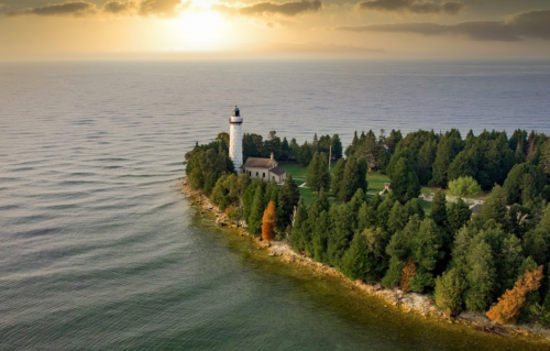 Aerial view of a lighthouse on a lush, green island surrounded by calm waters at sunset.