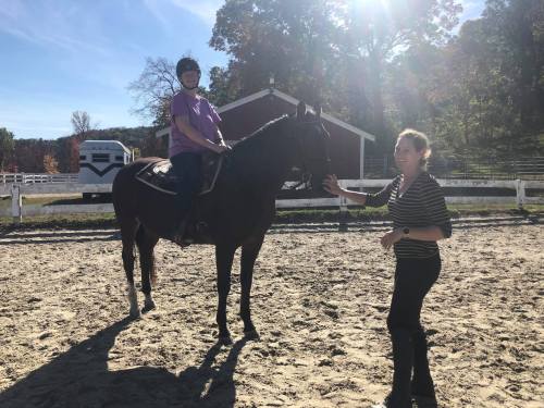 A child on a horse in a riding arena, with an adult standing beside them, both enjoying a sunny day outdoors.