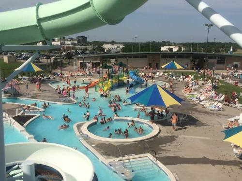 A busy water park with pools, slides, and colorful umbrellas, filled with people enjoying a sunny day.