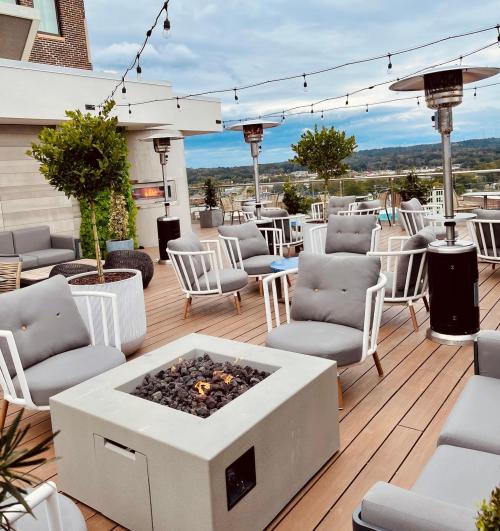 Rooftop lounge with modern seating, a fire pit, and patio heaters, surrounded by greenery and a scenic view.