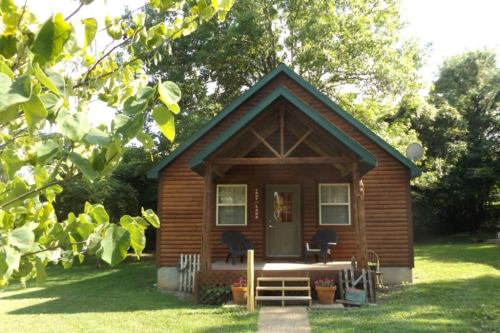 A cozy wooden cabin with a green roof, surrounded by trees and a well-kept lawn, featuring a front porch and flower pots.