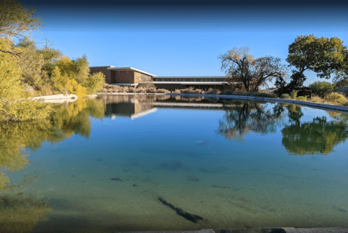 A calm, reflective pond surrounded by trees and a modern building under a clear blue sky.