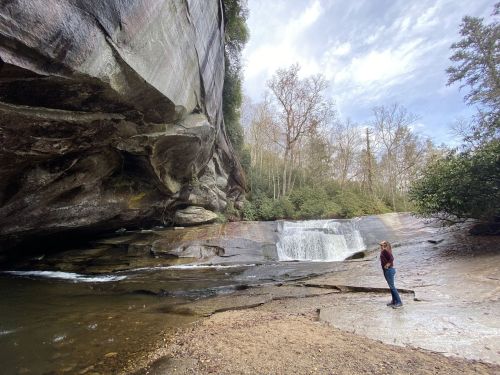 A person stands near a waterfall surrounded by rocky cliffs and lush greenery under a cloudy sky.