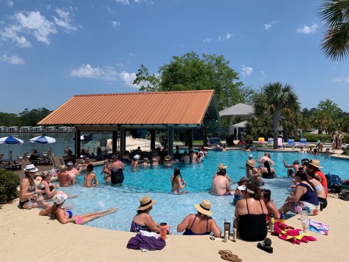 A crowded pool scene with people relaxing in the water and lounging around, surrounded by palm trees and blue skies.