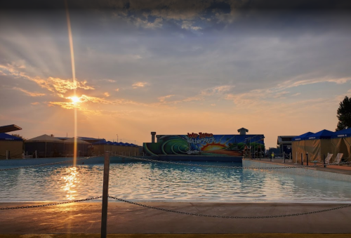 Sunset over a water park pool, with colorful murals and blue cabanas lining the edge. Calm water reflects the sky.