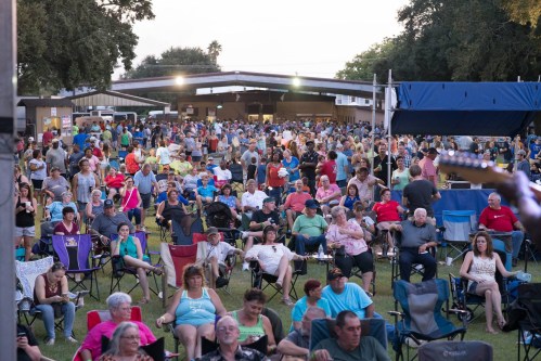 A large crowd of people sitting in chairs at an outdoor event, with a stage and trees in the background.