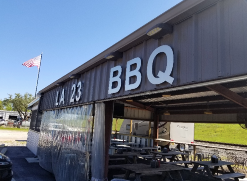 Exterior of LA 23 BBQ restaurant with a flag, featuring a metal structure and outdoor seating area.
