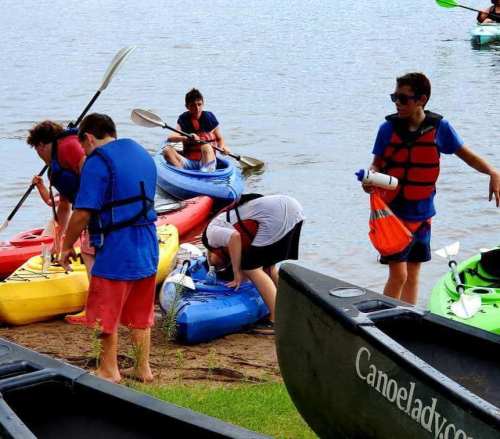 A group of people preparing kayaks by the water, wearing life jackets and gathering supplies on a sandy shore.