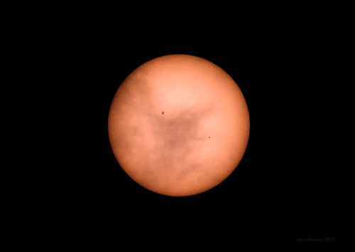 A close-up view of the sun with a hazy orange appearance and two small dark spots visible on its surface.