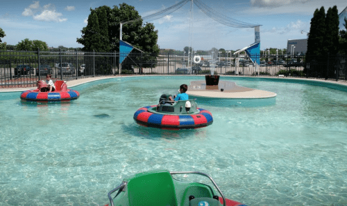 Two colorful bumper boats float in a clear pool under a blue sky, with trees and a fence in the background.
