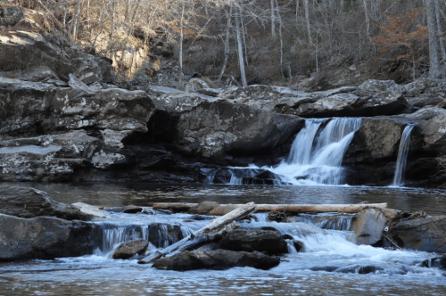 A serene waterfall cascades over rocks into a calm pool, surrounded by trees and natural scenery.