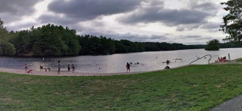 A panoramic view of a lake with people swimming and playing on the shore, surrounded by trees under a cloudy sky.