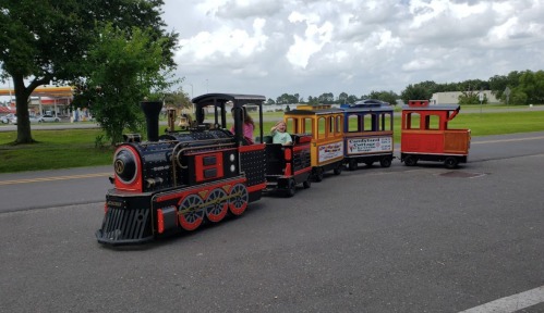 A colorful miniature train with several cars, parked on a road with trees and clouds in the background.