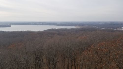 A panoramic view of a lake surrounded by bare trees and distant hills under a cloudy sky.