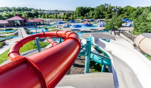 Aerial view of colorful water slides at a water park, with blue and white umbrellas and pools in the background.