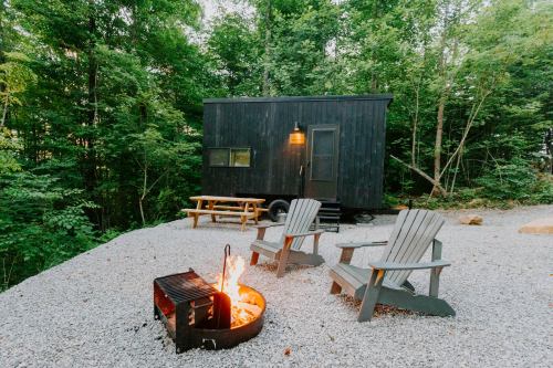 A cozy black tiny house surrounded by trees, with a fire pit and two chairs on a gravel area.
