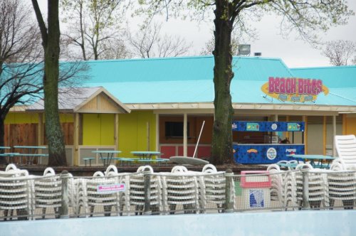 A colorful beach-themed concession stand with picnic tables and stacked chairs near a pool on a cloudy day.