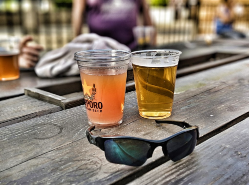 Two cups of beer on a wooden table, one light pink and one golden, with sunglasses nearby.