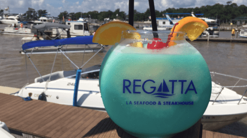 A colorful cocktail in a large glass with fruit garnishes, set against a marina with boats in the background.