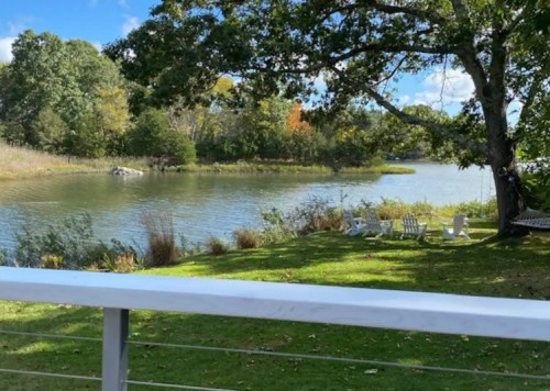 A serene riverside view with green grass, a few white chairs, and trees under a blue sky with scattered clouds.