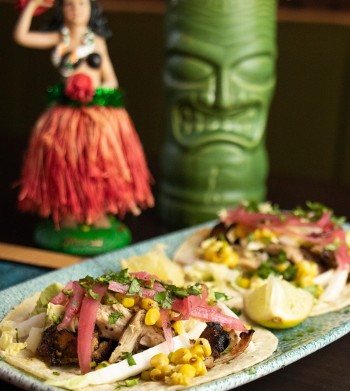 Two tacos filled with grilled chicken, corn, and toppings, with a tiki mug and hula figurine in the background.