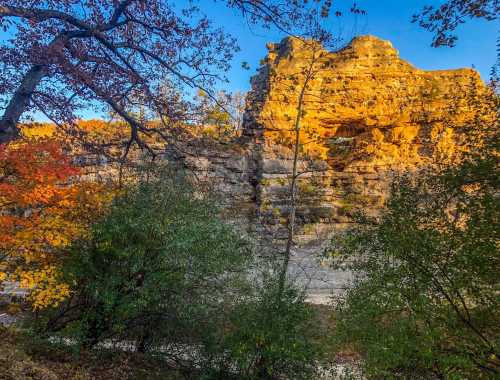 A rocky cliff illuminated by sunlight, surrounded by colorful autumn foliage and greenery.