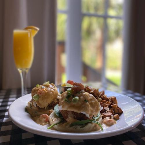A plate of eggs benedict with greens and hollandaise sauce, served with crispy potatoes and a mimosa in the background.