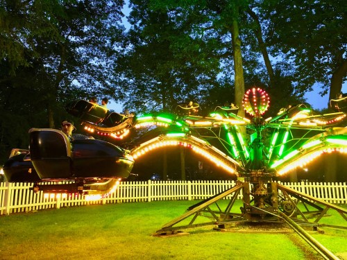A brightly lit amusement ride with green and yellow lights, set in a park surrounded by trees at dusk.