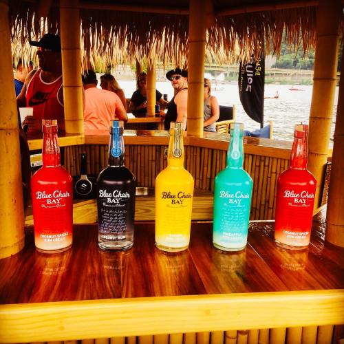 Five colorful bottles of Blue Chair Bay rum displayed on a wooden bar under a thatched roof, with people in the background.
