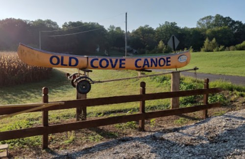 A yellow canoe sign reading "Old Cove Canoe" mounted on a wooden post near a grassy field and corn.