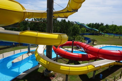Colorful water slides twist and turn over a sunny water park, surrounded by green trees and blue skies.