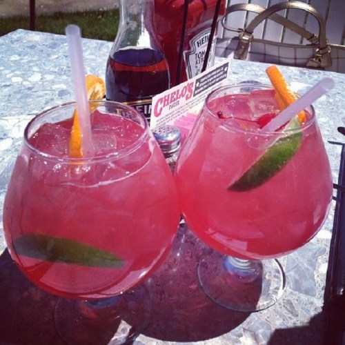 Two colorful cocktails in large glasses, garnished with lime and orange slices, on a table with condiments in the background.