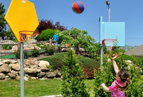 A child throws a basketball towards a hoop in a sunny outdoor setting with greenery and mini-golf in the background.