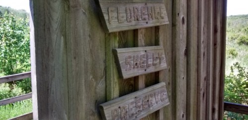 Wooden signs on a post display the names "Florence," "Shelby," and "Pittsville" against a natural backdrop.