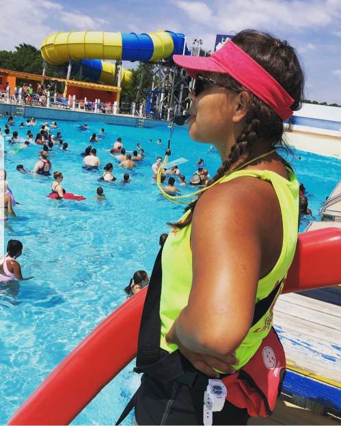 A lifeguard in a bright yellow shirt and pink visor stands by a pool, watching over swimmers enjoying the water.