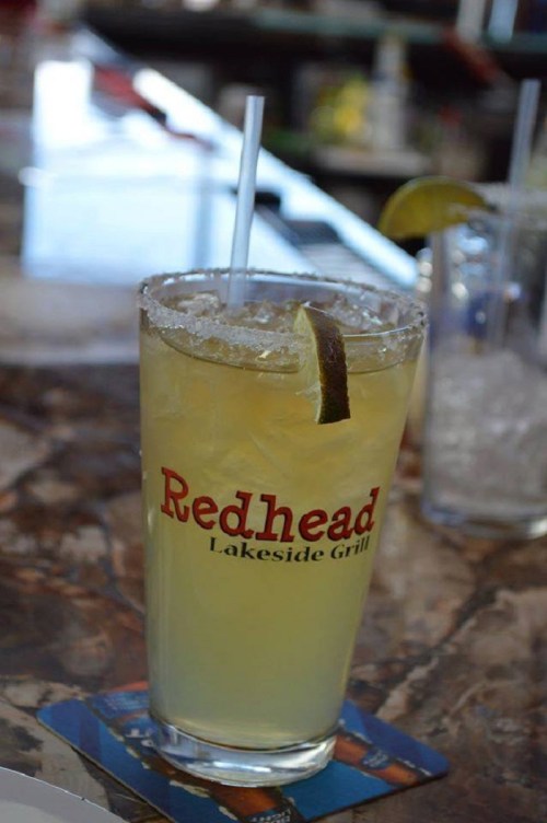 A glass of margarita with a lime wedge, featuring the logo of Redhead Lakeside Grill, on a bar countertop.