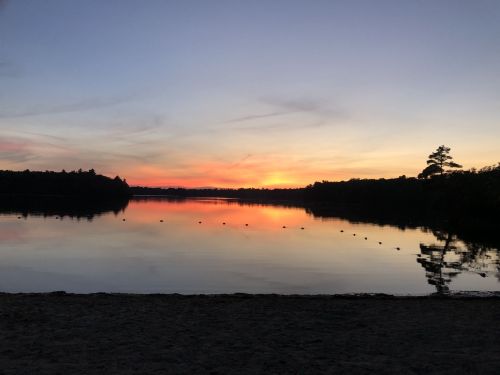 A serene lake at sunset, reflecting vibrant orange and purple hues in the water, surrounded by trees.