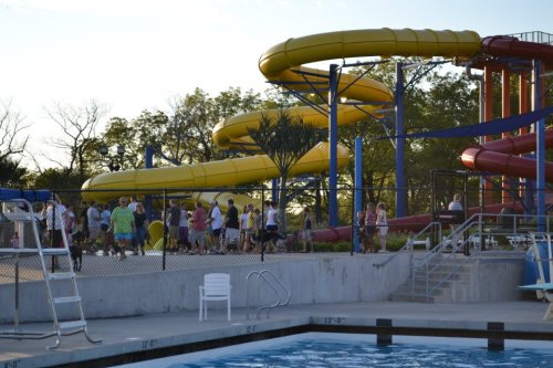 A water park scene with colorful slides, a swimming pool, and people walking near the slides on a sunny day.